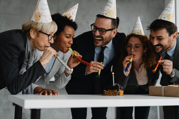 Group of business people in office celebrating birthday with cake, party hats and blowers. Corporate celebration, teamwork and cheerful workspace atmosphere