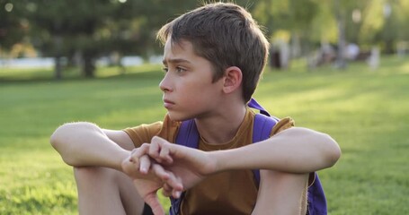 Thoughtful child boy with backpack sitting on green grass in park at sunset, hugging knees in concentrated pose.Calm and reflective mood, daylight, natural background, harmony with nature.Kid dreaming