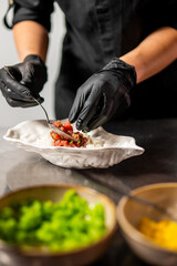 Chef in black gloves plating poke or tartare over rice in a leaf-shaped bowl. Fresh ingredients and vibrant colors highlight professional food preparation
