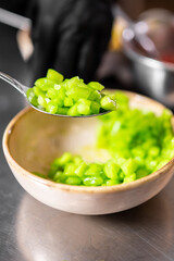 Close-up of gloved hand holding spoon with chopped green bell peppers over bowl. Food preparation in kitchen setting. Ingredient handling and cooking process concept.