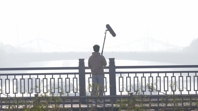 Sound recordist holding microphone in silhouette by riverside bridge in misty morning