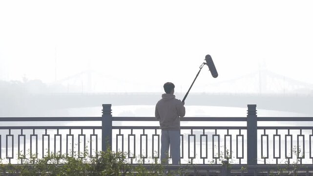 Sound recordist holding microphone in silhouette by riverside railing on foggy morning