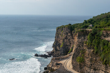 Dramatic cliffside view of the turquoise ocean at Uluwatu, Bali. Rugged coastline with waves crashing against the rocks and surfers in the distance