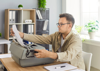 Writer typing at vintage typewriter in office workspace. A focused man at a desk inserts paper into a retro machine by notes and sheets in sunlight. Analog creativity and focused productivity.