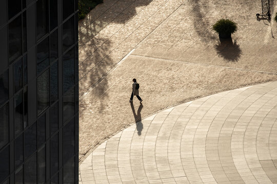 A solitary person walks through a sunlit urban plaza, casting a long shadow on the patterned ground. The scene captures architectural lines, open space, and minimalism in a city environment