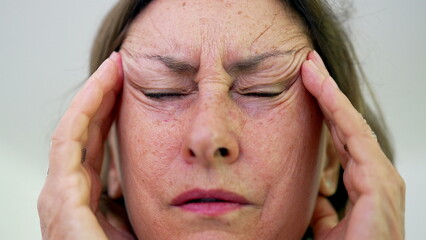 Close-up of senior woman with eyes tightly shut, pressing fingers to temples, intense focus on pain...