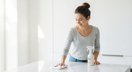 Young woman cleaning countertop with spray bottle and cloth in bright kitchen