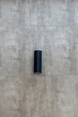 Minimalist black cylindrical outdoor wall lamp or sconce mounted on a modern grey concrete tile wall. Simple, clean architectural lighting detail