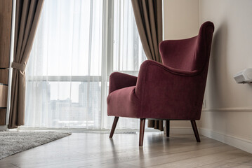 Modern interior with stylish burgundy armchair near window. Cozy living room design with curtains, wooden floor, and city view outside. Contemporary furniture for home comfort and relaxation.