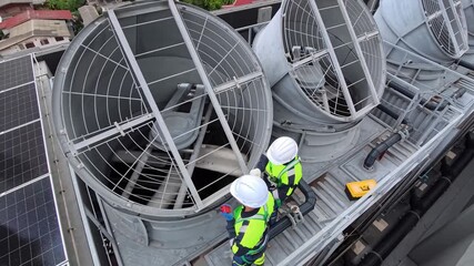 Two workers perform maintenance on large cooling fans situated on the rooftop of a commercial building in a bustling urban environment. They are dressed in safety gear and inspecting equipment.