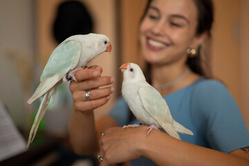 Smiling woman holding two parrots on her hand and arm indoors.
