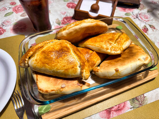 A beautiful glass dish holds several golden, homemade Chilean empanadas, perfectly arranged on a table next to a refreshing glass of soda, ready to be served.
