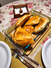 A close-up, top-down view of freshly baked Chilean empanadas, golden and delicious, served in a glass dish on a rustic wooden board with a floral tablecloth.