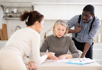 Fototapeta premium Thoughtful old woman compiling testament while middle-aged man and woman competing with each other in the kitchen