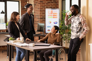 Asian female wheelchair user smiling and talking with her coworkers, emphasizing diversity and accessibility in the workplace. Multiethnic startup team having a cheerful conversation at the office.