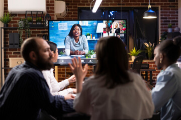 Multiethnic colleagues discussing startup project plans in late night meeting. A video conference with remote team is visible on digital screen, symbolizing modern business collaboration.