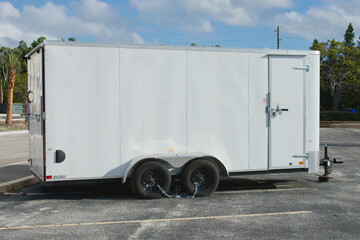 White Enclosed Cargo Trailer Parked in a Sunny Lot. Standing stationary in a sunny parking lot, secured by chains, under a clear sky background.

