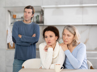 Fototapeta premium Man quarrels with his wife in kitchen, standing behind her, and elderly mother hugs and calms her daughter