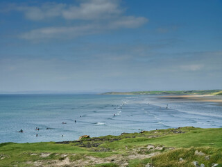 View from a cliff on a busy ocean with many surfers. Warm sunny day with blue cloudy sky. Popular sport and hobby. Bundoran, Ireland.