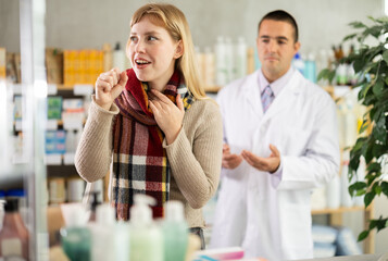 Young woman with a cold throat and a wrapped scarf chooses cough medicines and antipyretics against the background of a pharmacist in a pharmacy. Buying antiviral medications