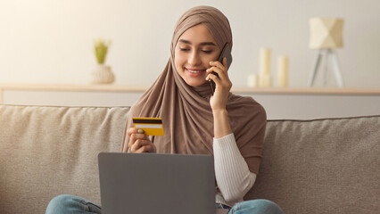 Online payment at home. Successful woman doing online shopping using computer. Smiling middle eastern pretty lady in hijab speaks on phone and holds credit card in living room interior, free space