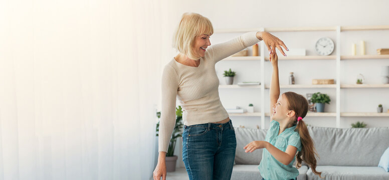 Happy mature lady and casual little daughter dancing waltz at home, standing in living room, full length photo. Loving grandmother teaching granddaughter how to dance having fun together at home - Powered by Adobe