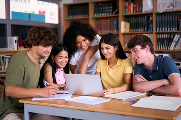 Five diverse high school students are smiling and working together on a project in the library, using a laptop and taking notes, demonstrating teamwork and collaboration in education