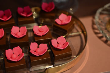 flowers shaped chocolates, 
gourmet chocolate bonbons with pink flower decorations, elegant dark chocolate cubes for a special event, artisanal confectionery on a decorative golden tray