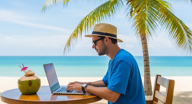 Man working on laptop at beachside table with coconut drink under a palm tree on a sunny day