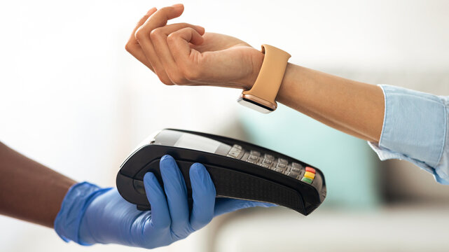 Cropped close up view of female client paying with smart watch on payment terminal, black man in protective gloves holding pos machine in hand. Digital contactless cashless transaction concept