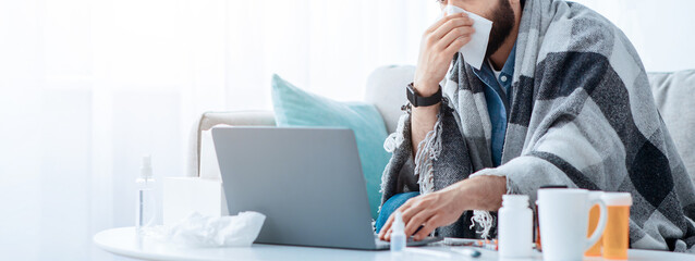 Ill arab guy sneezing and blowing runny nose in paper tissue, working distantly on laptop computer,...