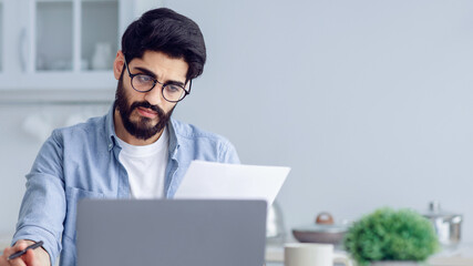 Stressed arab man calculating home budget, sitting in kitchen, copy space. Young man suffering...
