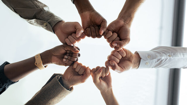 Success And Teamwork Concept. Below view of young diverse group of business people in protective medical masks making fist bump standing in circle. Workers do fist pump together celebrating good deal - Powered by Adobe