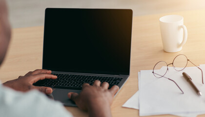 Fototapeta premium Work with website, send email, video call and search in internet at home or office. Middle aged african american male typing on laptop with empty screen on desk with cup and glasses, indoors, close up