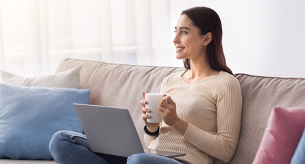 Naklejka premium Enjoying free time and social networks at home. Beautiful young muslim woman work on laptop and drinking coffee. Cheerful pensive arab lady on sofa looking towards empty space in living room interior
