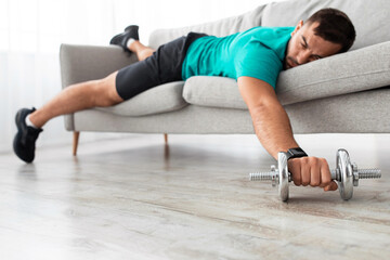 Tired millennial male with beard lies on sofa with dumbbells rest after exercise in living room interior. Relax, take break and pause at training, body care, workout at home during covid-19 pandemic