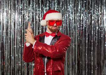 Man dressed in red Santa suit and sunglasses, playful posing like secret agent in front of shimmering silver background. Christmas and New Year Holiday celebration, party invitations concept 
