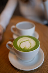 Matcha latte art forming a heart shape in a white cup resting on a wooden table, with another cup of coffee softly blurred in the background, creating a cozy cafe atmosphere