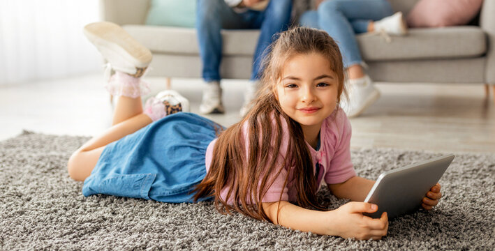 Leisure time. Cute little girl using digital tablet, lying on floor carpet and smiling at camera. Young family spending time at home, parents relaxing on sofa with laptop, selective focus on child