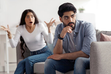 Fototapeta premium Quarrel, scandal at home, conflict, relationship problems, divorce. Angry excited young arab woman yelling and gesturing at offended husband, ignoring her in living room interior, free space