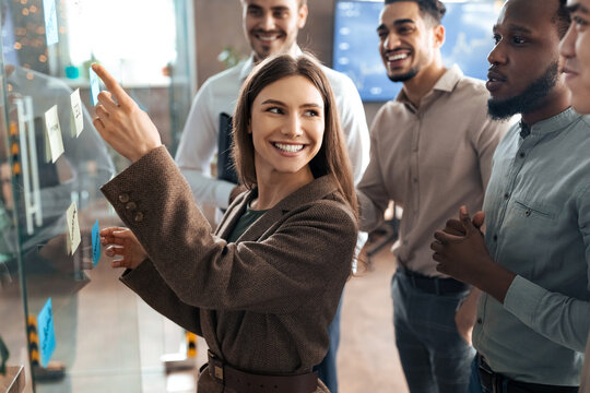 Teamwork Concept. Creative group of smiling professional businesspeople brainstorming, wonan pointing at sticky post-it paper notes on glass wall, discussing ideas analysing new business strategy