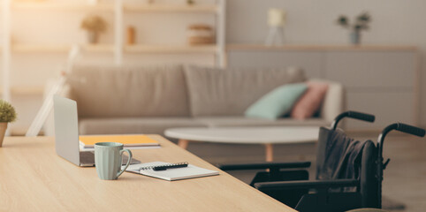 Workplace for disabled person, remote job at home during covid-19 quarantine. Wheelchair with nobody, wooden table with cup, notepad and laptop, sofa, furniture in living room interior, copy space