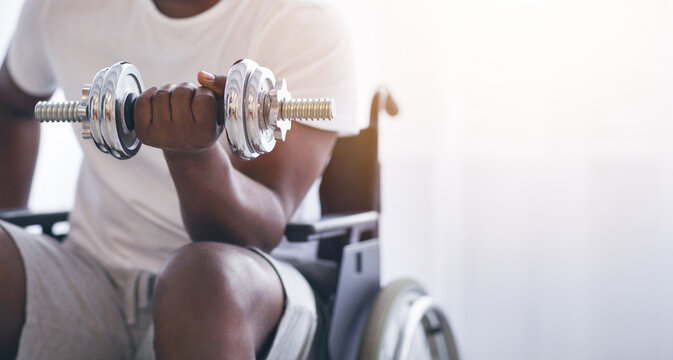 Man exercising at home and recovering health care, self-isolation and sport. Smiling millennial african american male disabled in wheelchair raises dumbbell on window background in interior, cropped - Powered by Adobe