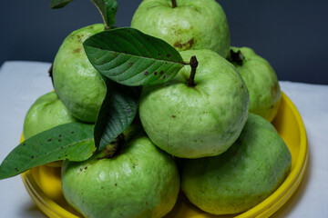 Close-up A pile of fresh, ripe green guavas and their leaves, served on a yellow plate against a clean gray background.
