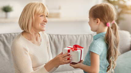 Celebrating Concept. Portrait of cute little girl making surprise for her grandma holding and giving present box, greeting woman with holiday. Granddaughter congratulating smiling mature lady