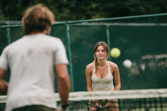 A female tennis player stands on a sunny court, eyes fixed on an approaching ball as a blurred opponent signals a rally behind a chain link fence.