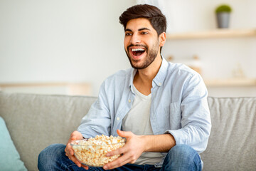 Joyful arab man watching his favorite movie on tv and eating popcorn, relaxing on couch in living room. Happy eastern guy resting on comfortable sofa, enjoying weekend, having domestic leisure