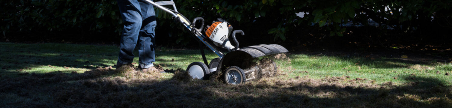 Senior man operating a gas powered dethatching machine, tossing up pieces of dead lawn thatch and moss, grass lawn care and maintenance, dramatic background of sunlight and shade
