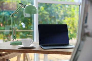 Home workspace. Laptop, lamp and cup of drink on wooden desk near window indoors