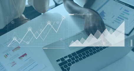 Two business women reviewing data at office desk with laptop, report, tablet and graph overlays